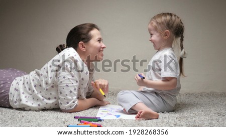 Mother, nanny, teaches child girl to draw. Happy family playing together at home on floor. Mom helps her daughter learn to draw on paper by coloring with multi-colored pencils and felt-tip pens.