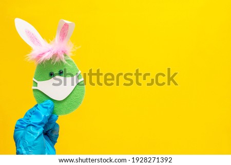 Hand in medical glove holds coronavirus easter painted green egg wearing medical mask with bunny ears Background yellow Illuminating