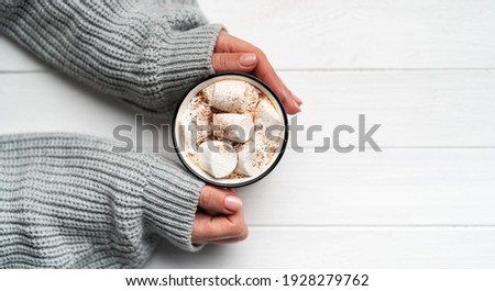 Woman holding cacao mug filled with marshmallow in hands on white wooden table background, top view