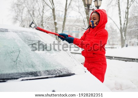 African american woman in red hoodie and face mask clean car from snow in winter day.