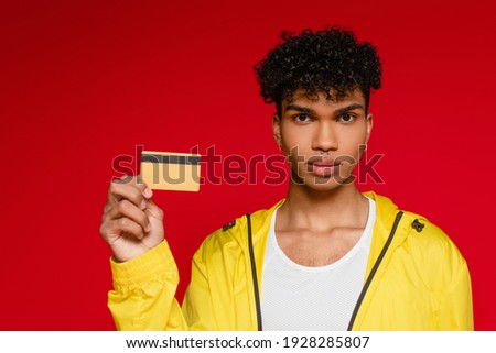 pierced african american man in jacket holding credit card isolated on red