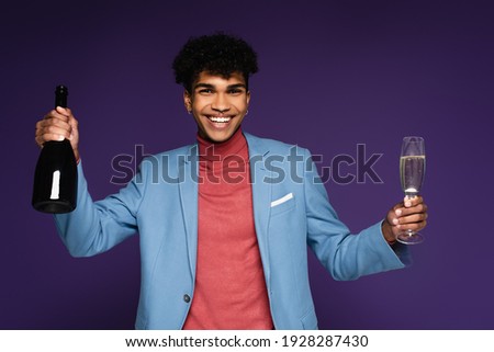 african american man in blue blazer holding glass and champagne bottle on purple