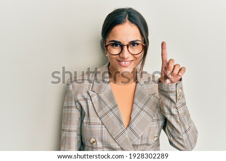 Young brunette woman wearing business jacket and glasses showing and pointing up with finger number one while smiling confident and happy. 