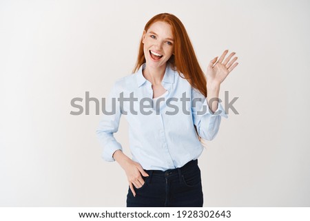 Redhead female hr manager saying hello, waving hand and smiling friendly, greeting you, standing over white background. Royalty-Free Stock Photo #1928302643