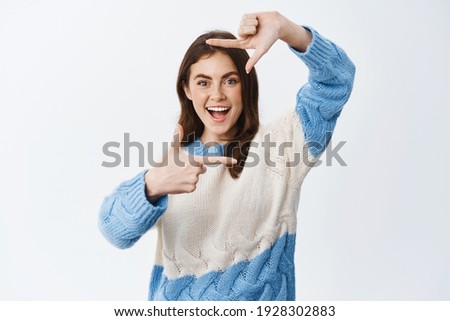 Excited smiling woman looking through hand camera frames with amazed face expression, capturing moment, standing in sweater against white background.