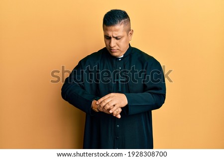 Young latin priest man standing over yellow background checking the time on wrist watch, relaxed and confident 