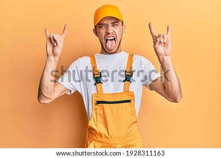 Hispanic young man wearing handyman uniform shouting with crazy expression doing rock symbol with hands up. music star. heavy music concept. 