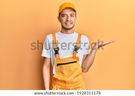 Hispanic young man wearing handyman uniform smiling cheerful presenting and pointing with palm of hand looking at the camera. 