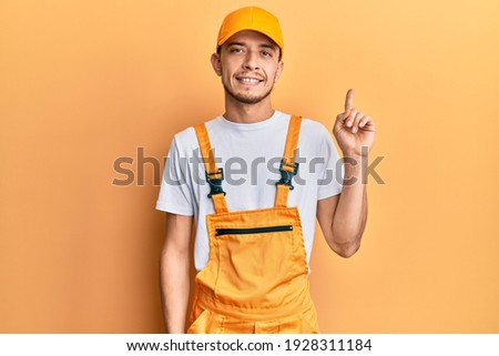 Hispanic young man wearing handyman uniform with a big smile on face, pointing with hand finger to the side looking at the camera. 