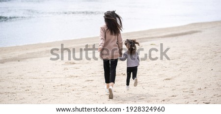 Rear view photography of cute white baby girl and her young mother running together happily on spring sandy beach. Family dressed in sweaters or jumpers of pastel pink and purple colors