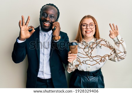 Young interracial couple using smartphone and drinking a cup of coffee doing ok sign with fingers, smiling friendly gesturing excellent symbol 