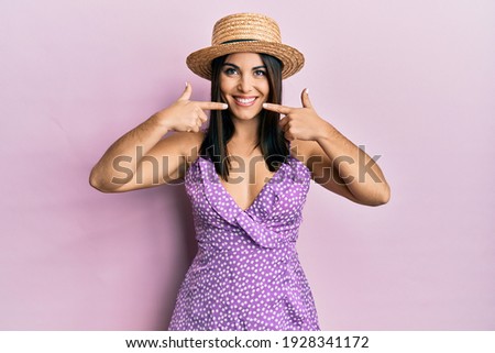 Young brunette woman wearing summer dress and hat smiling cheerful showing and pointing with fingers teeth and mouth. dental health concept. 