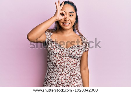Young latin woman wearing casual clothes doing ok gesture with hand smiling, eye looking through fingers with happy face. 