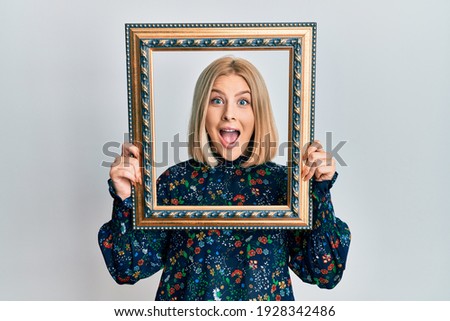 Young blonde woman holding empty frame celebrating crazy and amazed for success with open eyes screaming excited. 