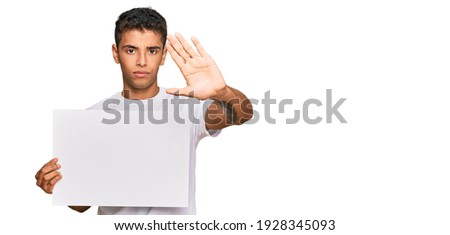 Young handsome african american man holding blank empty banner with open hand doing stop sign with serious and confident expression, defense gesture 