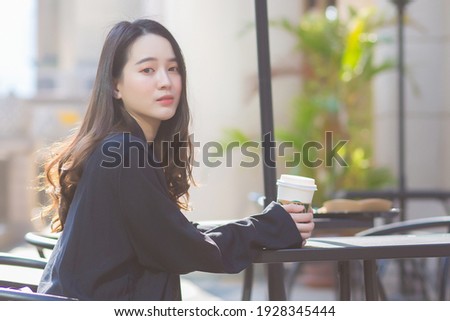 A beautiful Asian woman in a dark blue suit is sitting on a chair looking out. In front of a coffee shop in a sunny morning.
