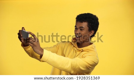cheerful african american teenage boy in hoodie taking selfie isolated on yellow