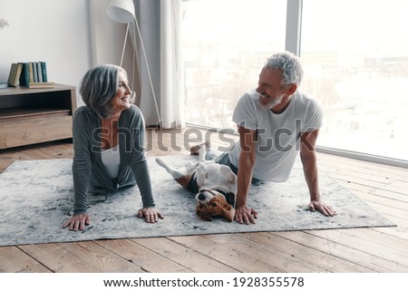 Loving senior couple in sports clothing doing yoga and smiling while spending time at home with their dog Royalty-Free Stock Photo #1928355578