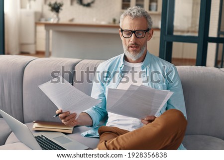 Thoughtful senior man checking the papers with serious face while sitting on the sofa at home Royalty-Free Stock Photo #1928356838