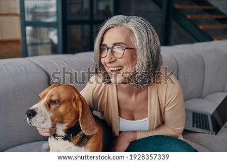 Beautiful senior woman in casual clothing spending time with her dog while sitting on the sofa at home Royalty-Free Stock Photo #1928357339