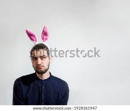 Caucasian man with pink hare ears is dressed in blue stylish shirt and looks sideways with caution and suspicion. Empty space for your text or ad on white background. Man with ears of Easter bunny.