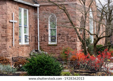 The front side of a stone church building surrounded with plants and trees.