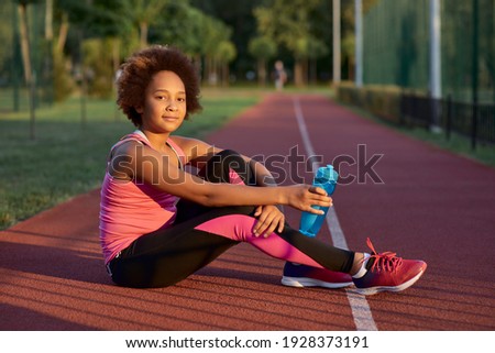 Sporty little girl with bottle of water sitting on running track