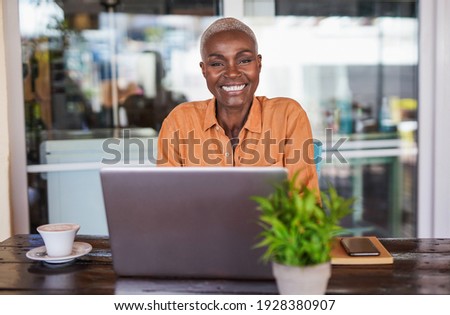 Beautiful mature african woman sitting in a bar outdoor and working with laptop - Black woman looking in camera and smiling Royalty-Free Stock Photo #1928380907
