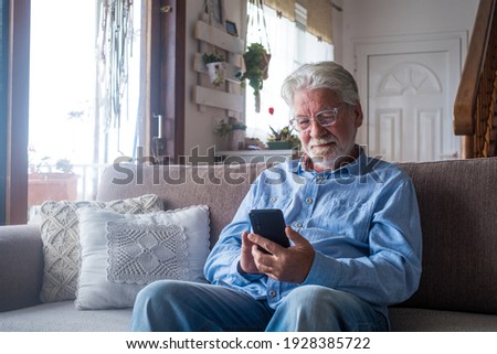 Old man smiling sitting on the sofa in the living room holding phone, enjoying using smartphone feeling satisfied sending messages, calling friends, surfing web online concept Royalty-Free Stock Photo #1928385722