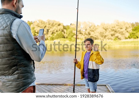 family, generation, summer holidays and people concept - father with smartphone photographing his happy smiling son with fishing rod showing thumbs up on river