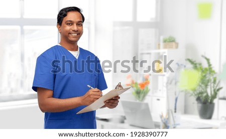 healthcare, profession and medicine concept - happy smiling indian doctor or male nurse in blue uniform writing report on clipboard over medical office at hospital background
