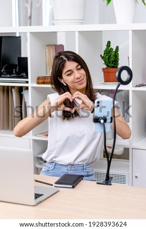 Mixed race influencer girl blogger  talking smartphone,live recording video blog on social network at home.The girl shows the heart symbol with her hands.Social media live streaming concept.