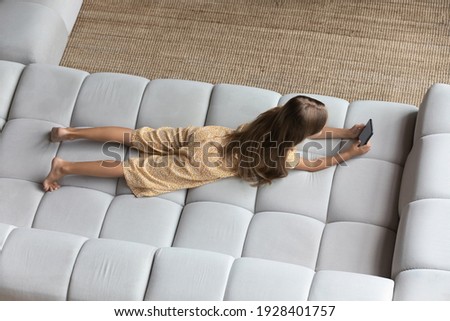 Funny pastime. Top above view of smart little girl lying on belly on comfortable couch at home spend free time using cell. Junior school age kid surf web safe after setting parental control on gadget
