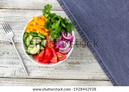 Plate of rainbow salad with different vegetables and herbs in white bowl with blue napkin and fork on white wooden background. Top view