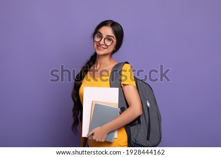 Education Concept. Portrait of smiling indian female student wearing eyeglasses, holding notebooks looking at camera. Woman wearing backpack posing isolated over purple studio background