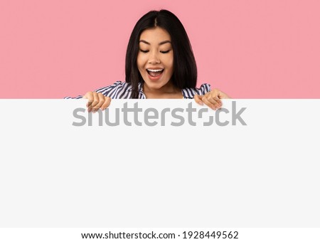 Wow. Excited asian female standing behind white empty board for advertisement over pink background. Cheerful lady with open mouth looking down at big blank advertisement placard, empty space for text