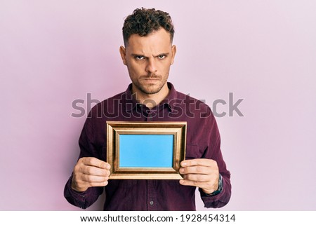 Young hispanic man holding empty frame skeptic and nervous, frowning upset because of problem. negative person. 
