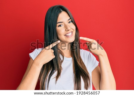 Young brunette woman wearing casual white tshirt over red background smiling cheerful showing and pointing with fingers teeth and mouth. dental health concept. 