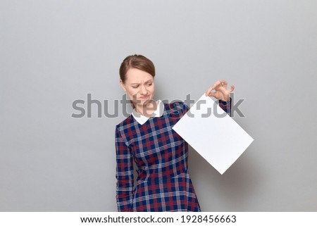 Studio portrait of disappointed young blond woman wearing checkered dress, holding paper sheet by the corner with displeasure, being dissatisfied with document, standing over gray background