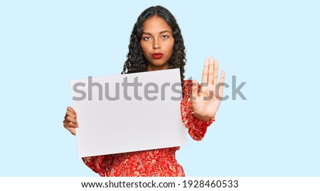 Young african american girl holding blank empty banner with open hand doing stop sign with serious and confident expression, defense gesture 