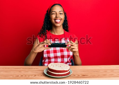Young african american girl wearing baker apron making carrot cake picture sticking tongue out happy with funny expression. 