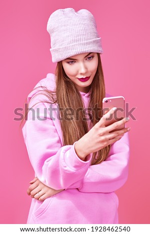 Modern teen girl in a pink sweatshirt and a hat takes a selfie on the phone on a pink background. Social networks concept. Youth generation. 