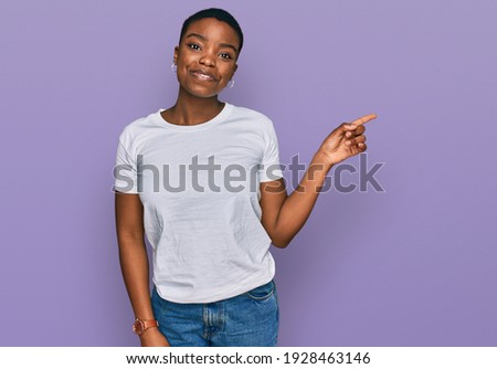 Young african american woman wearing casual white t shirt with a big smile on face, pointing with hand finger to the side looking at the camera. 