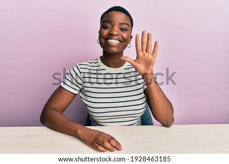 Young african american woman wearing casual clothes sitting on the table showing and pointing up with fingers number five while smiling confident and happy. 