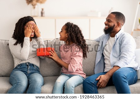Portrait of African American girl making surprise for her mum, giving present, greeting woman with Mother's day or birthday. Smiling lady covering eyes sitting on the couch in living room