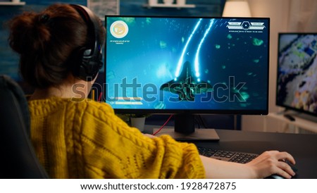 Pro gamer playing in the space shooter on personal computer. Woman gaming with teammates through headphones during online eSport tournament in action shot using technology network wireless Royalty-Free Stock Photo #1928472875