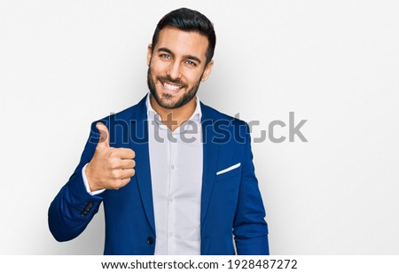 Young hispanic man wearing business jacket doing happy thumbs up gesture with hand. approving expression looking at the camera showing success. 