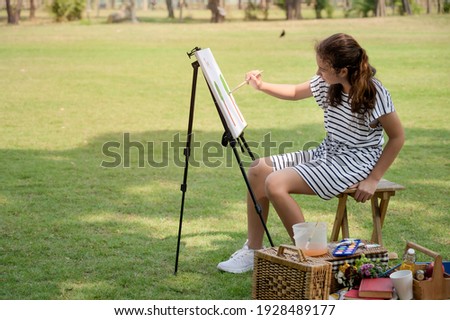 A half-Thai-European girl  is sitting on the wooden bench and painted on the canvas placed on a drawing stand, Be a part of learning outside of the school in the nature park