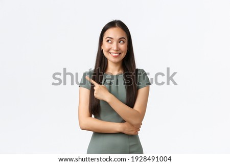 Small business owners, women entrepreneurs concept. Intrigued happy asian female in dress found excellent choice, pointing finger left and looking interested, smiling satisfied, white background Royalty-Free Stock Photo #1928491004