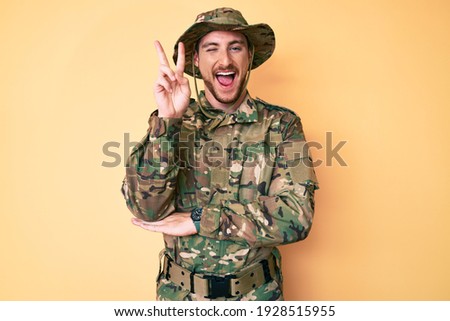 Young caucasian man wearing camouflage army uniform smiling with happy face winking at the camera doing victory sign with fingers. number two. 